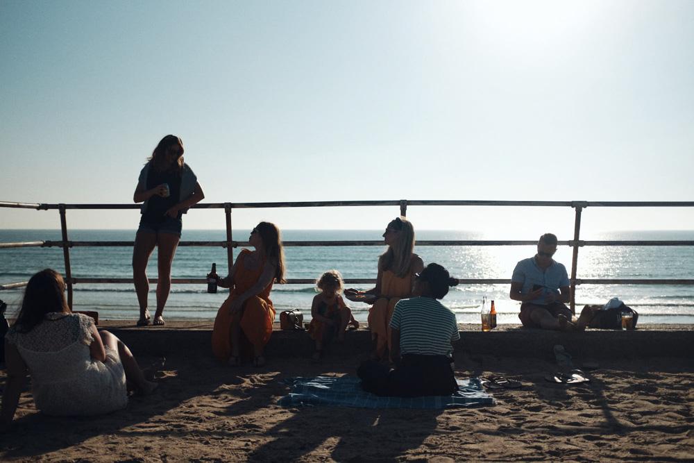 A group of people, mostly women and children, sit on a beach boardwalk with the ocean in the background. They appear to be enjoying a warm, sunny day, possibly with some drinks.