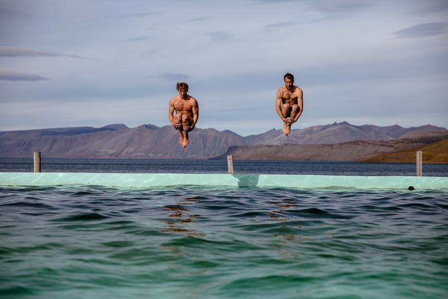 Two men, both naked, are in mid-air, jumping off a diving board into a large, blue, ocean-like pool, with a mountainous, rocky, and green landscape in the background.