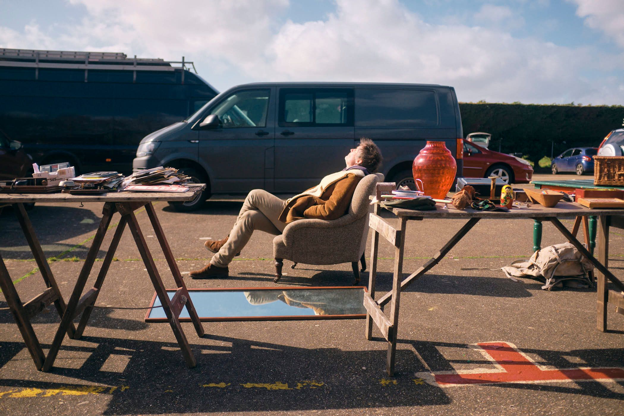 A man is sitting in a chair, surrounded by tables of antiques and furniture, in a parking lot. He is relaxed, with his eyes closed, and appears to be enjoying the sun.