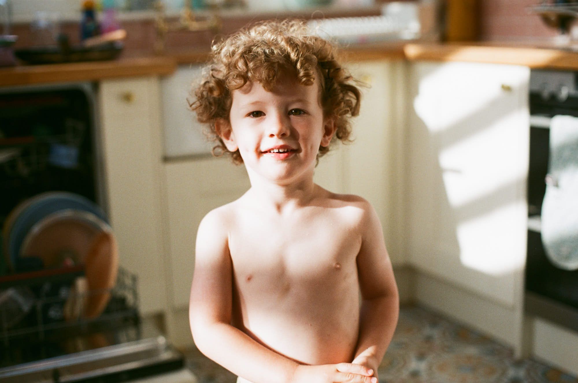 A young boy with curly hair and a big smile stands in a kitchen, possibly in front of a sink or counter. He appears to be enjoying himself, perhaps playing or helping with a task.