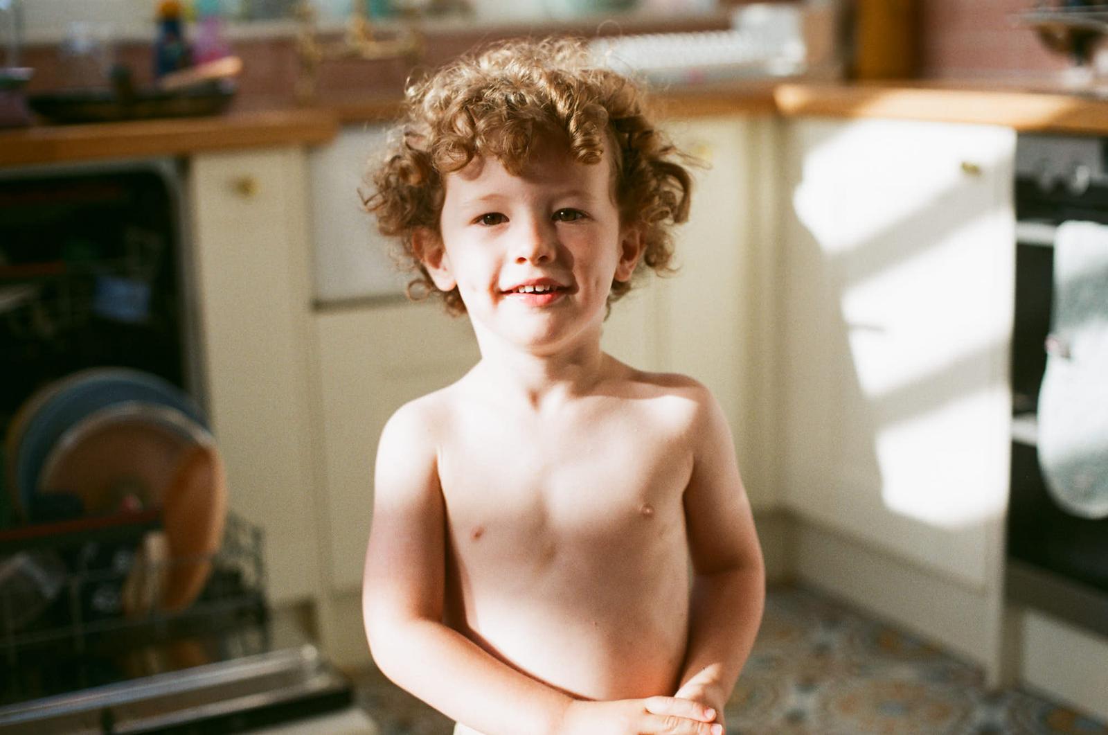 A young boy with curly hair and a big smile stands in a kitchen, possibly in front of a sink or counter. He appears to be enjoying himself, perhaps playing or helping with a task.