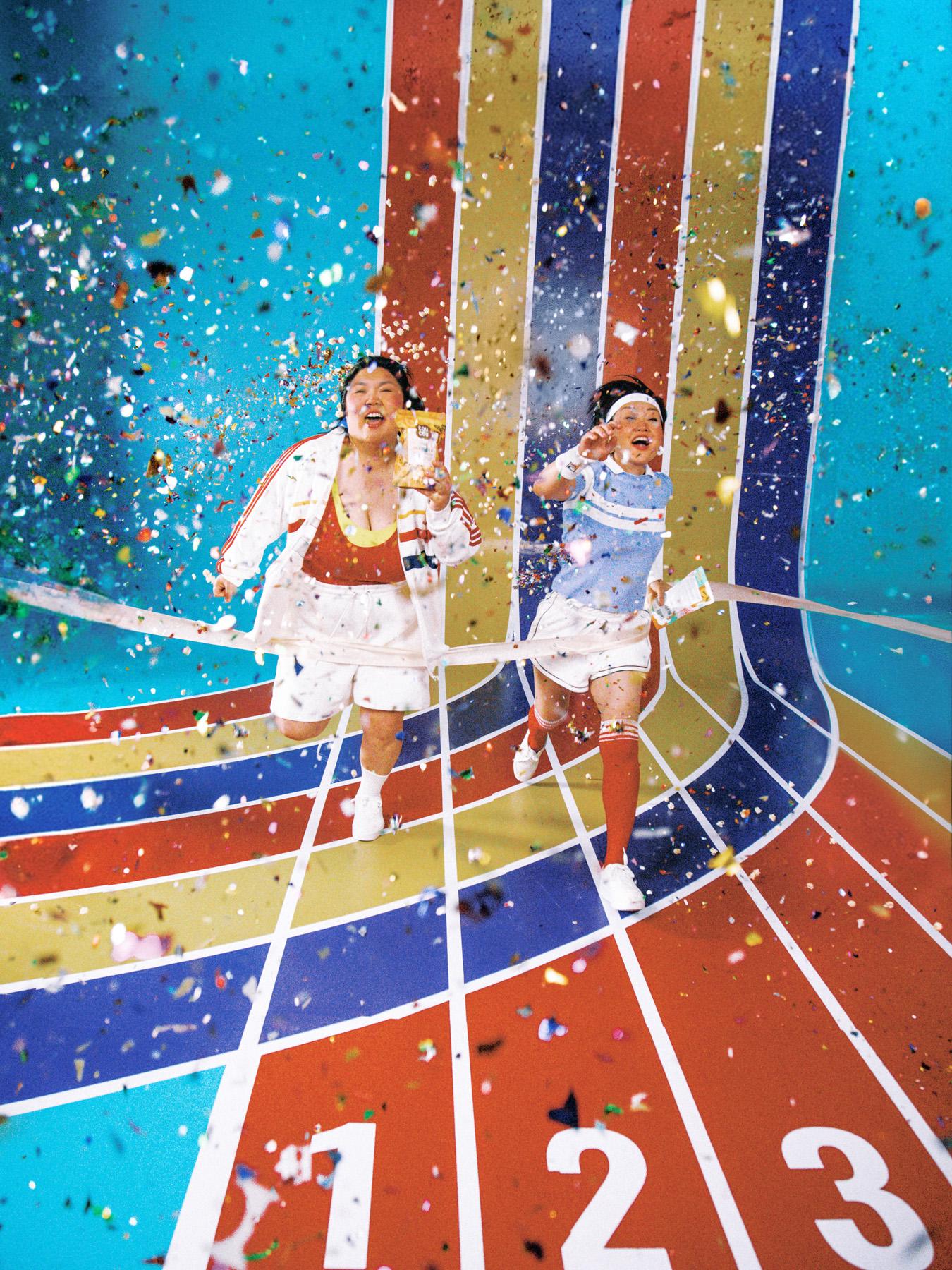 Two women in 1980s-inspired athletic wear running on a colorful, striped track, surrounded by confetti and celebratory atmosphere.