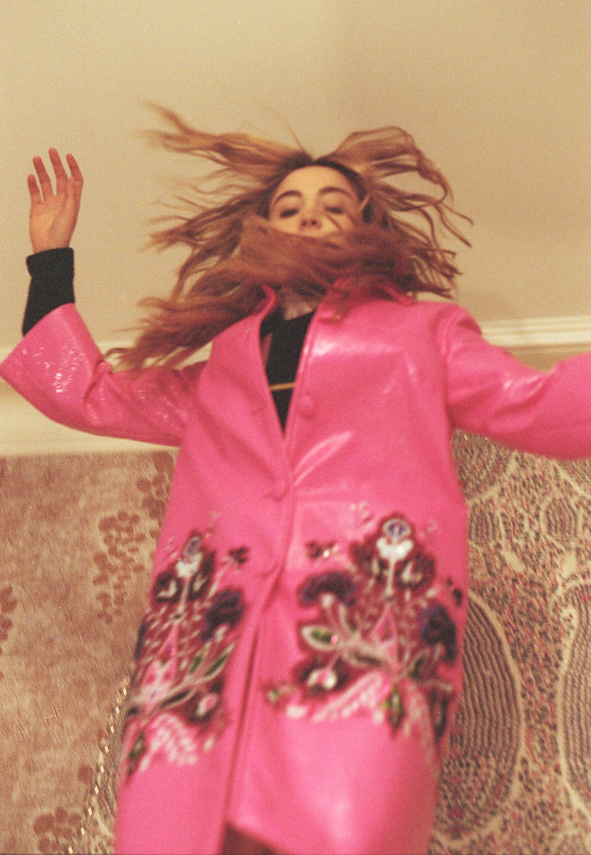 A woman lies on her back with limbs spread, wearing a pink, shiny, flaky coat with a floral pattern.