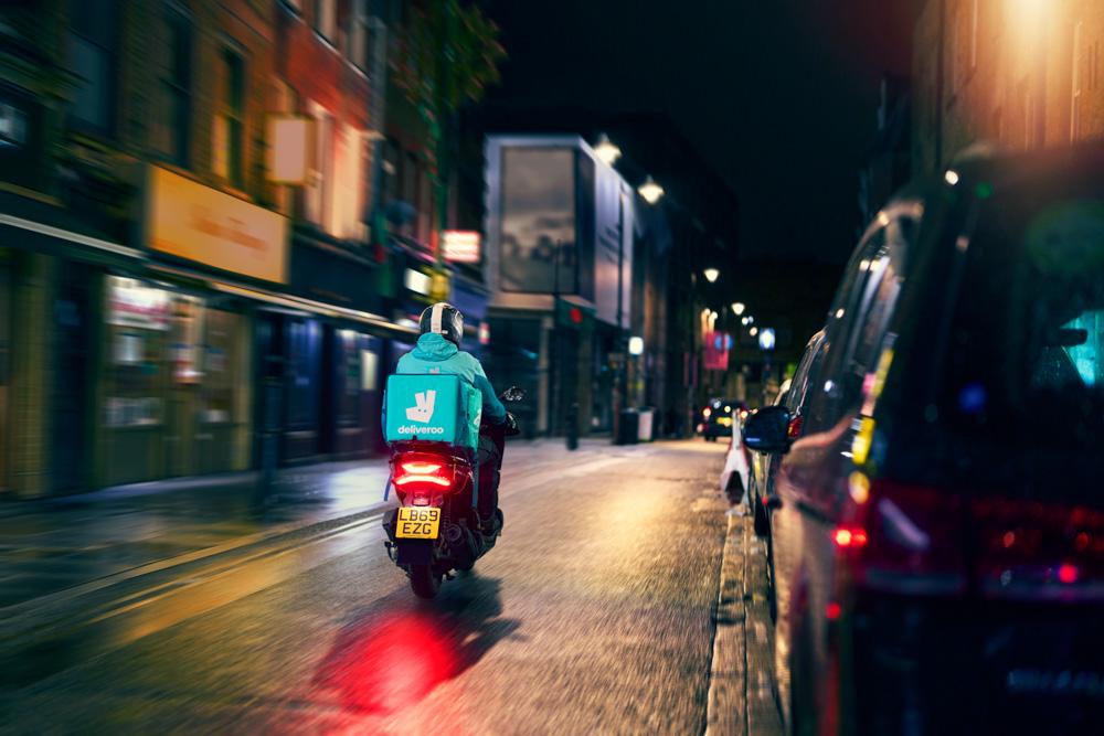 A person in a blue Deliveroo uniform riding a motorbike through a city street at night, with a Deliveroo food box on the back of the bike.
