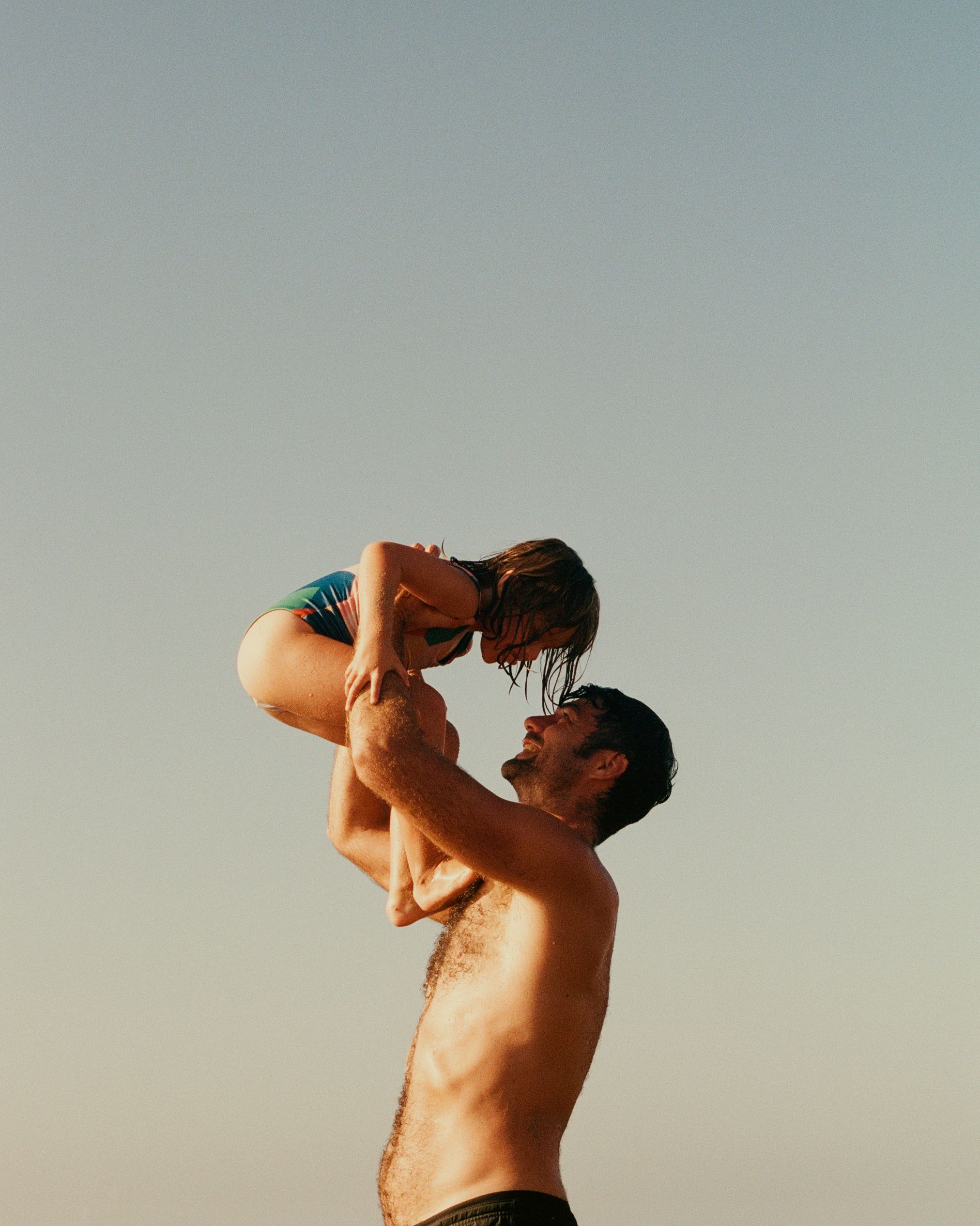 A man lifting a young girl above his head, both wearing swimsuits, on a beach or poolside.