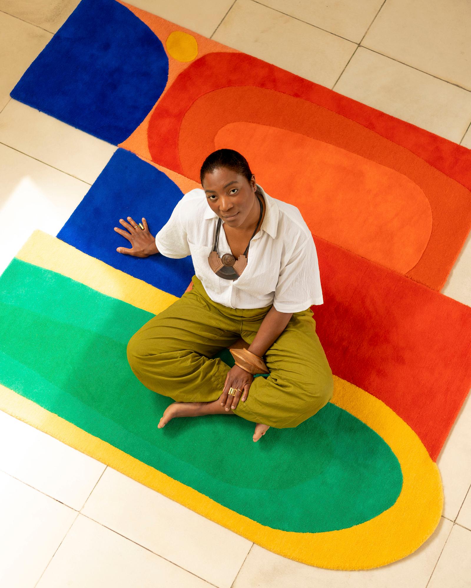 A woman with short hair and a white shirt sitting on a colorful rug, surrounded by various art pieces and sculptures.