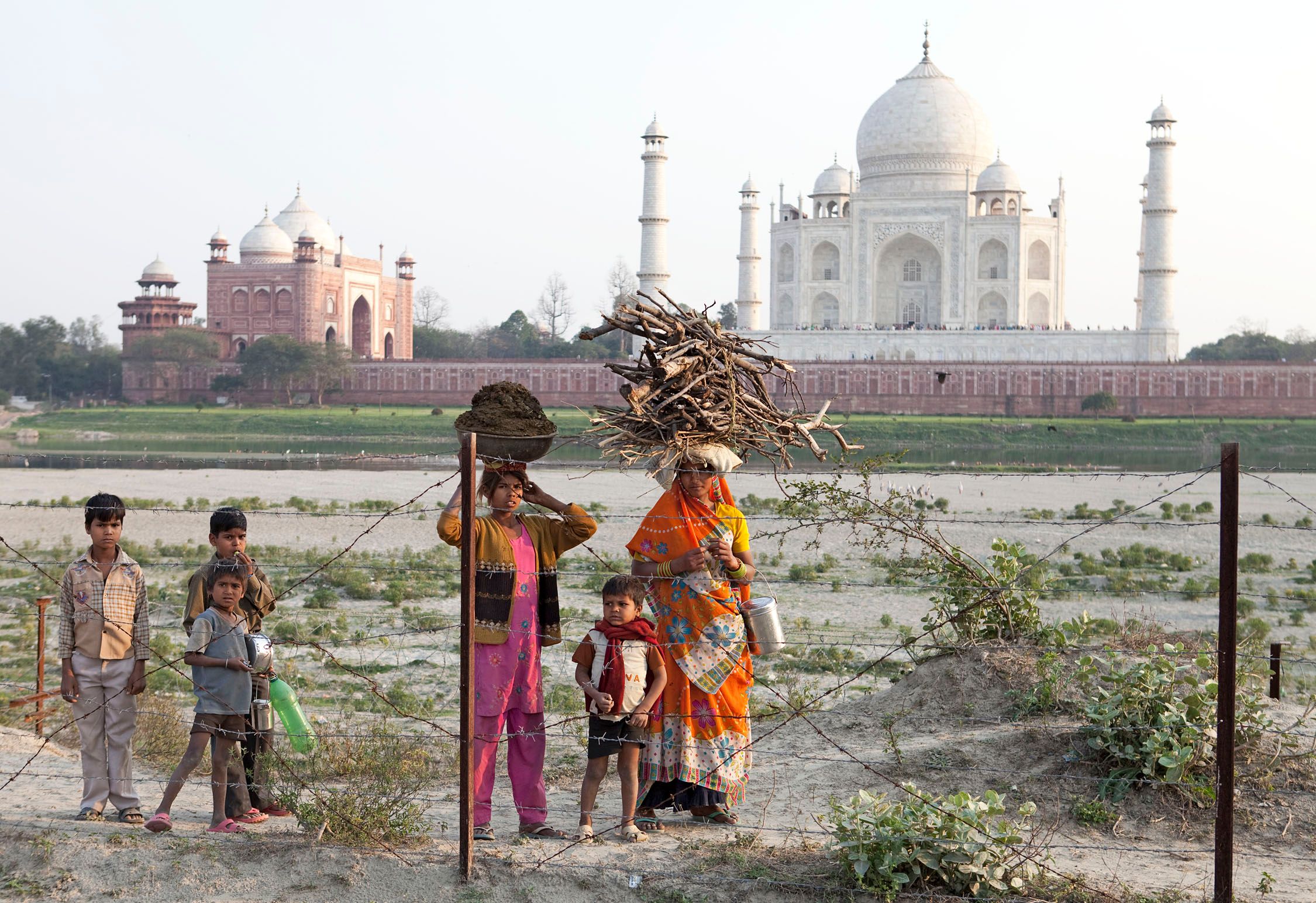 A group of people, including children and adults, standing in front of the Taj Mahal in Agra, India, with the monument's white marble domes and minarets visible in the background.