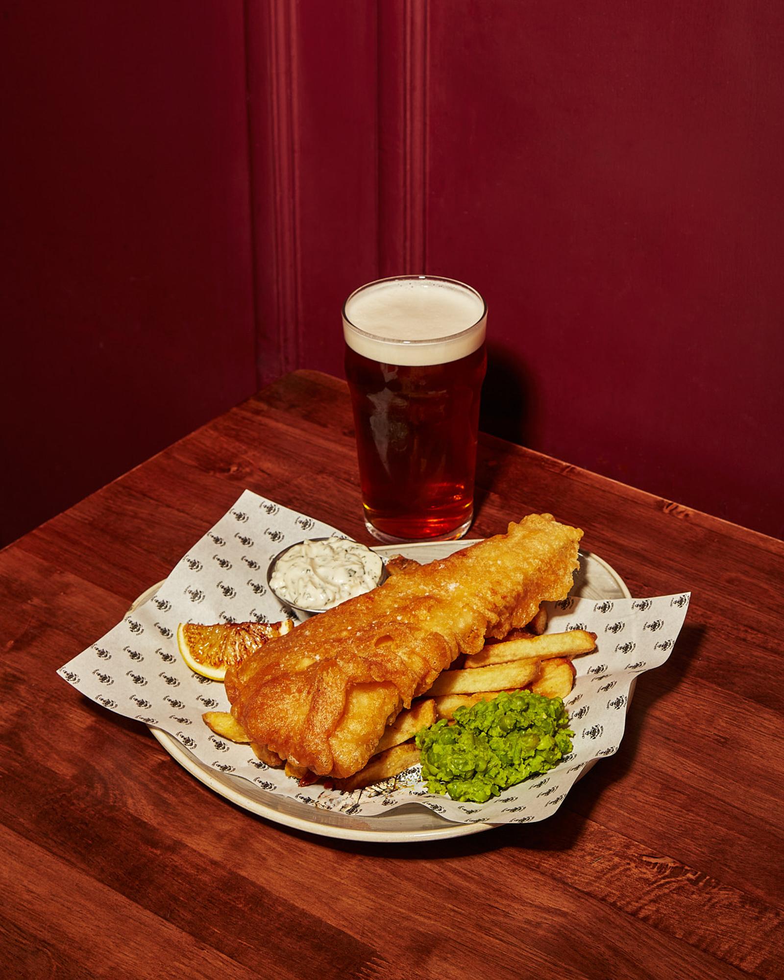 A plate of fish and chips, with a glass of beer, on a wooden table in a dimly lit, red-walled room.