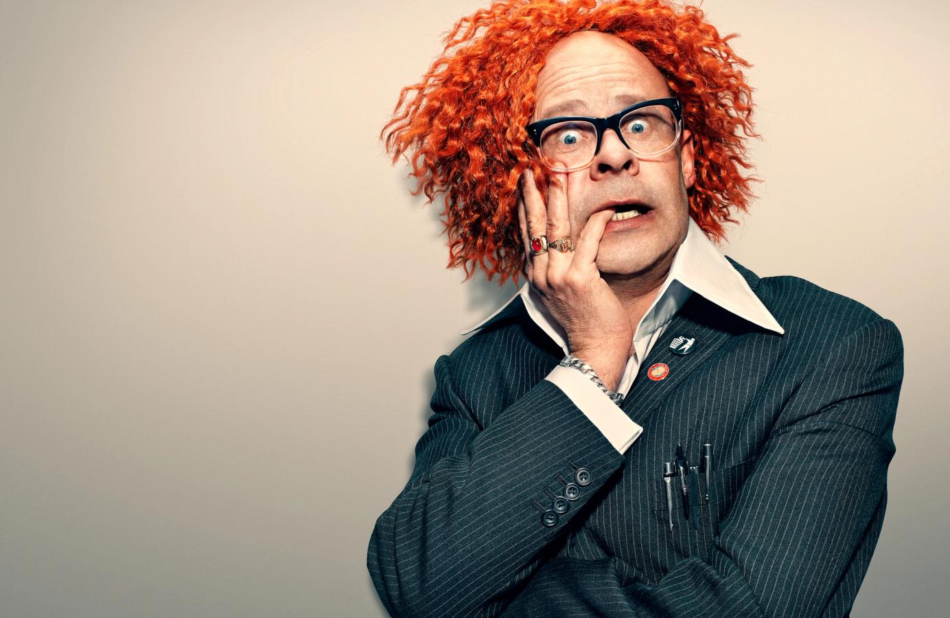 A man with curly red hair wears a dark pinstripe suit, white shirt, and red tie, has a red button on his lapel, and a pen in his breast pocket, looking surprised at the camera.