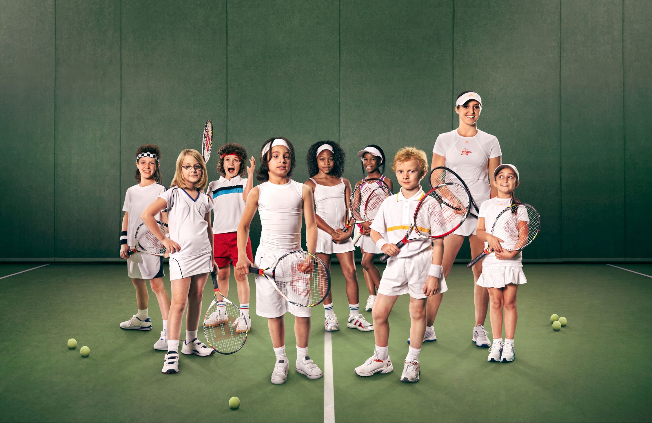 A group of children and a woman in tennis outfits, standing on a tennis court with tennis rackets and balls.