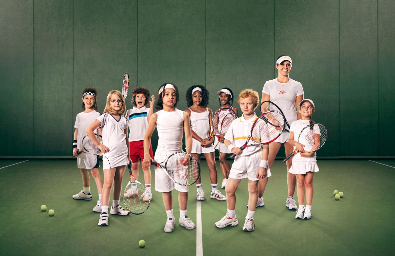 A group of children and a woman in tennis outfits, standing on a tennis court with tennis rackets and balls.