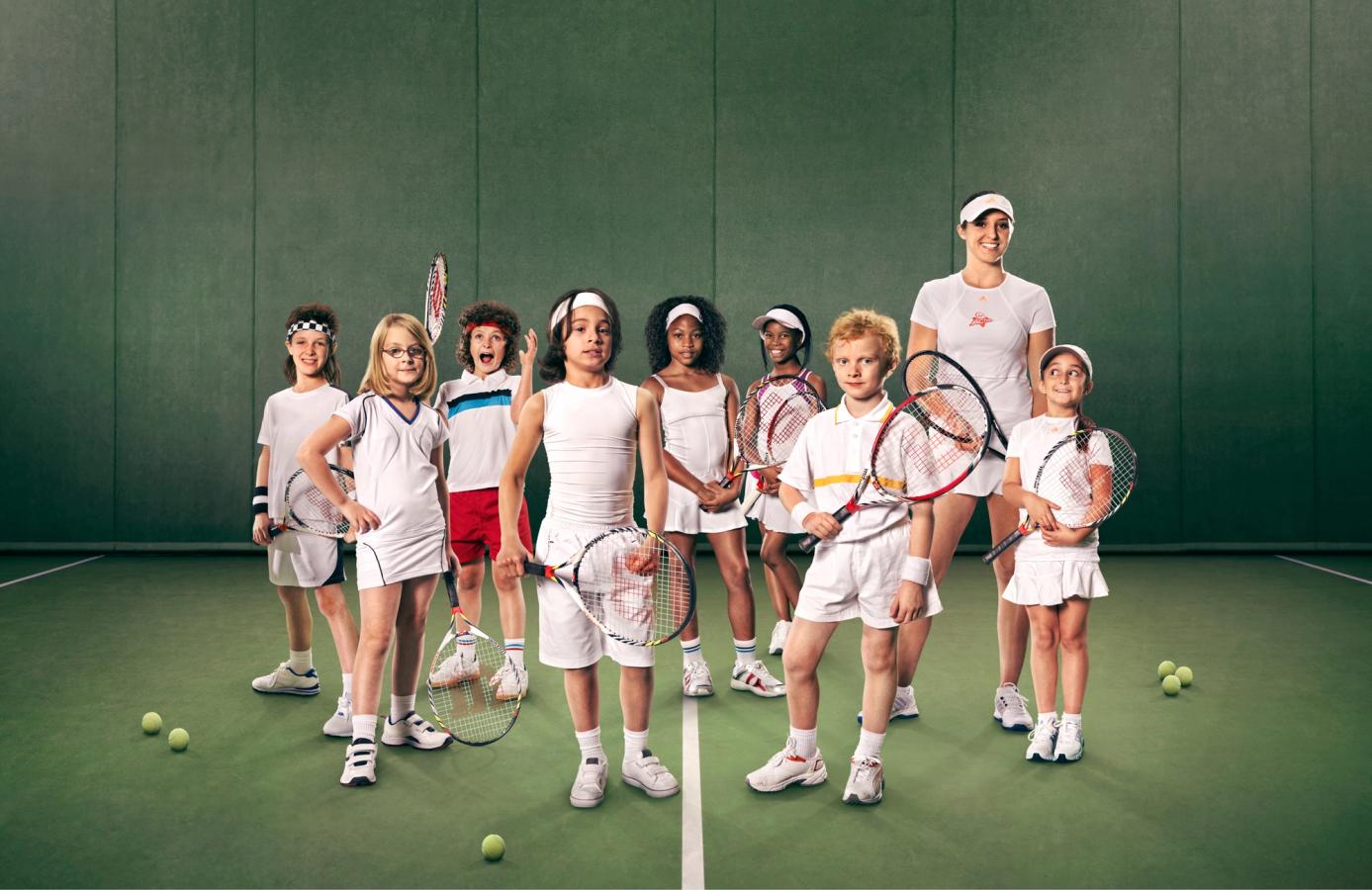 A group of children and a woman in tennis outfits, standing on a tennis court with tennis rackets and balls.