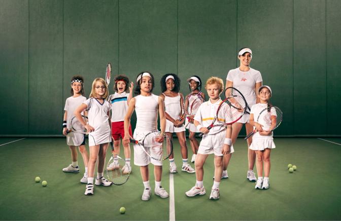 A group of children and a woman in tennis outfits, standing on a tennis court with tennis rackets and balls.
