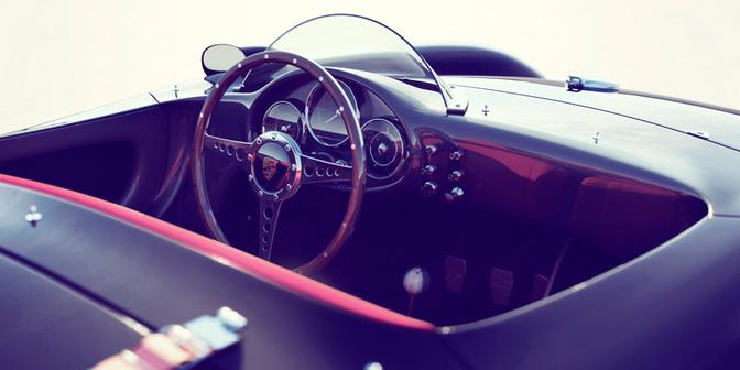 A close-up of a car's steering wheel and dashboard, with a focus on the leather-wrapped steering wheel and the iconic Porsche logo on the dashboard.