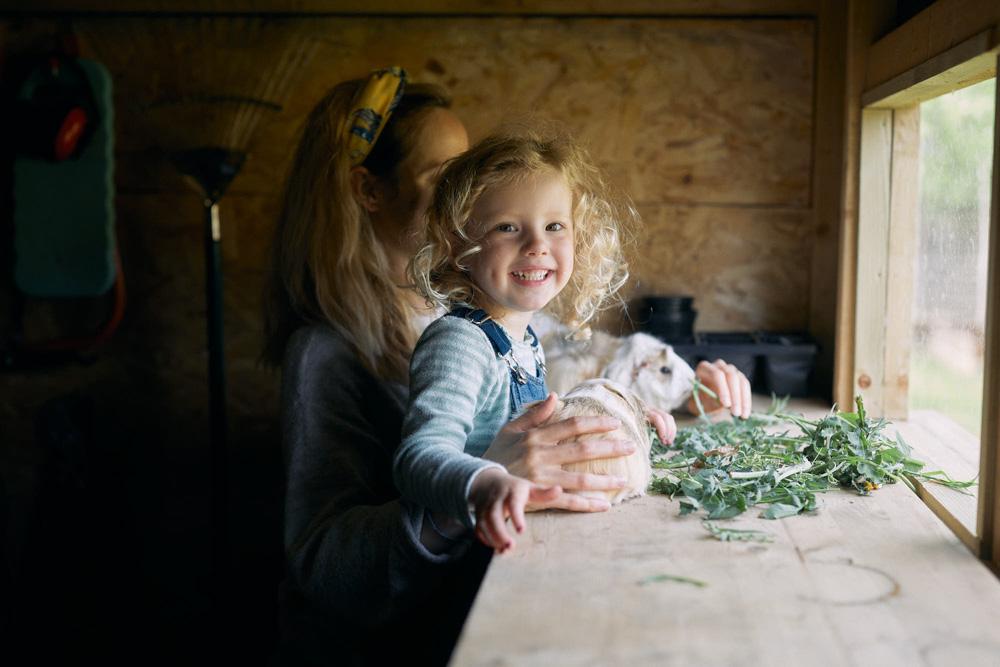 A young girl with curly blonde hair and her mother, both with similar features, are feeding a guinea pig on a table by a window. The girl is smiling and holding the guinea pig,.