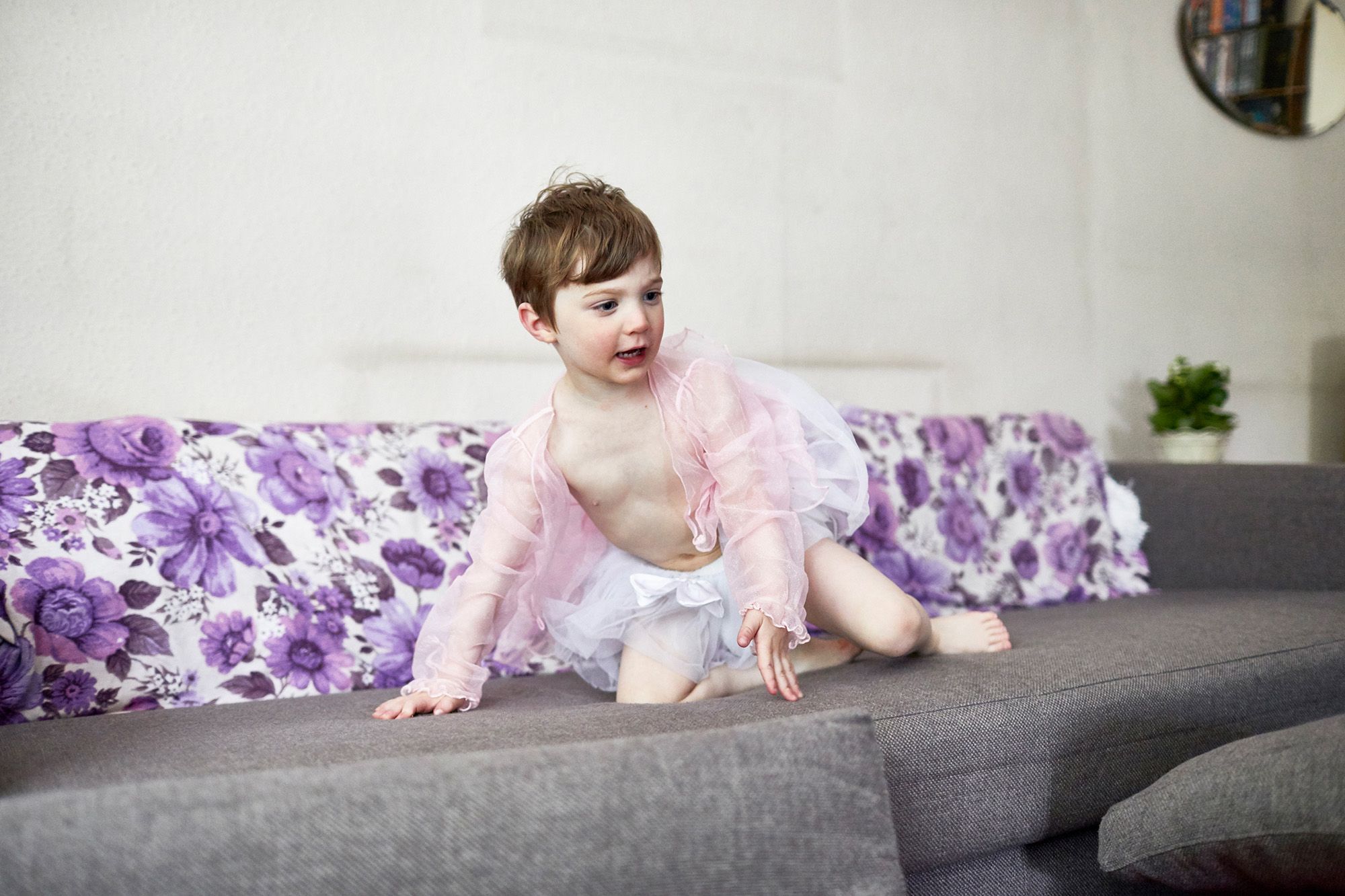 A young boy, likely a toddler, is sitting on a couch, wearing a pink tutu and a matching pink top, with a big smile on his face.