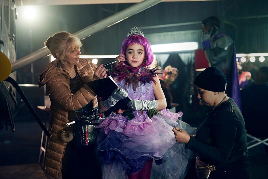 Two women in brown coats and two children in purple dresses and tiaras are on a film set.