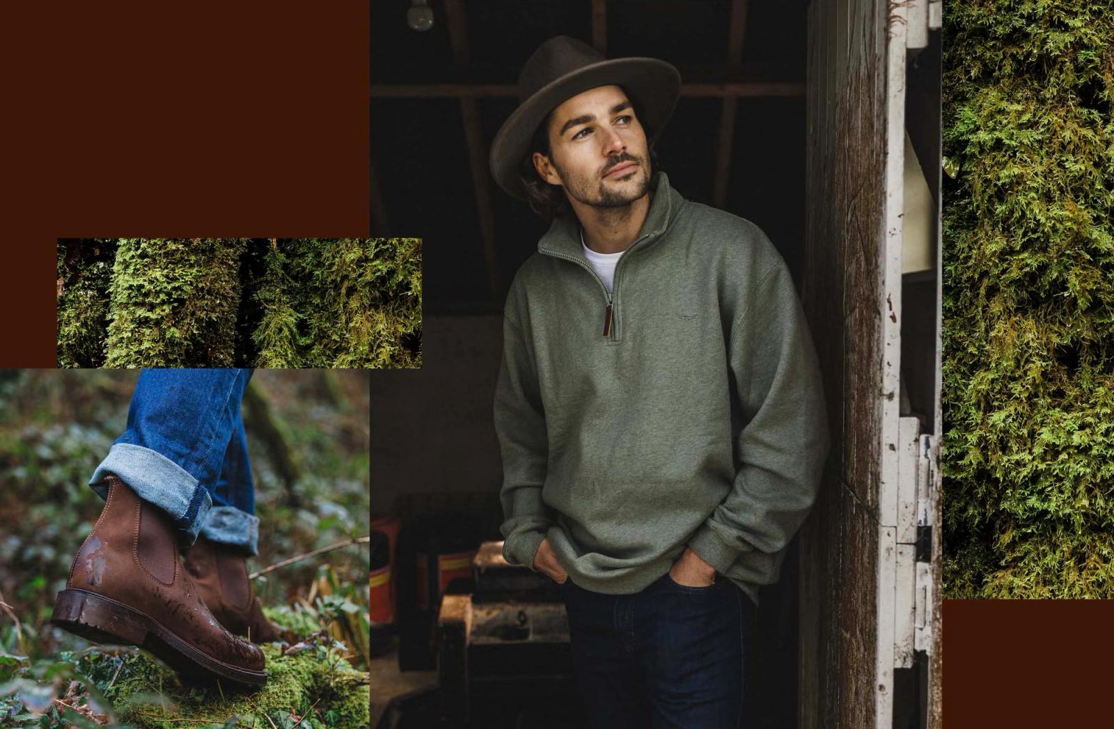 A man wearing a green quarter-zip sweater, jeans, and a hat, standing in a dark, moss-covered environment, with a focus on his clothing and surroundings.