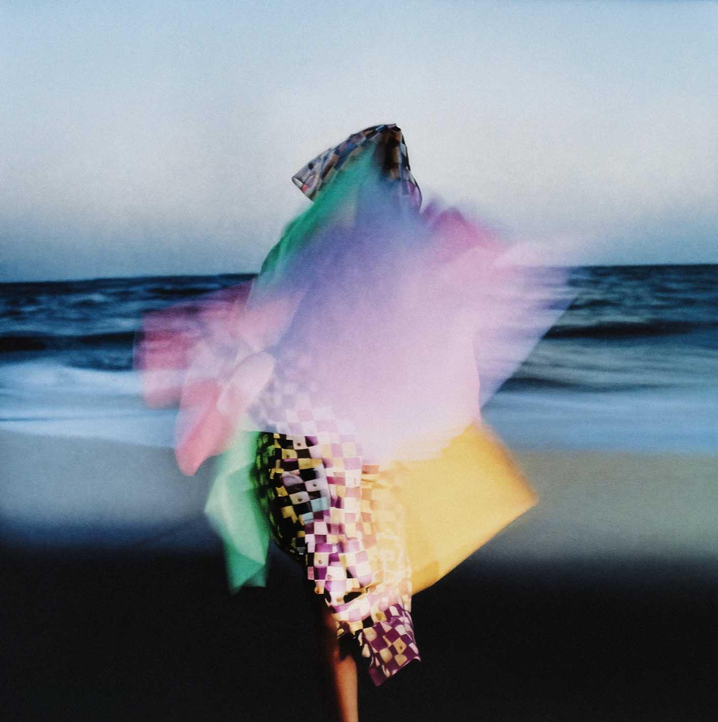 A person in a colorful, pixelated, 8-bit style outfit, standing on a beach with the ocean in the background.