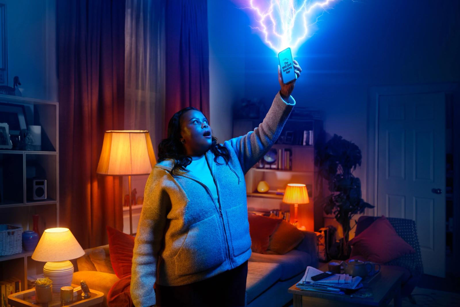 The image shows a woman standing in a living room with dark blue walls and warm lighting from several lamps. She is wearing a grey zip-up hoodie and is looking up in awe at a smartphone she is holding high in the air. The phone displays "The NHS Quit Smoking App" and has bright blue lightning bolts emitting from the top.