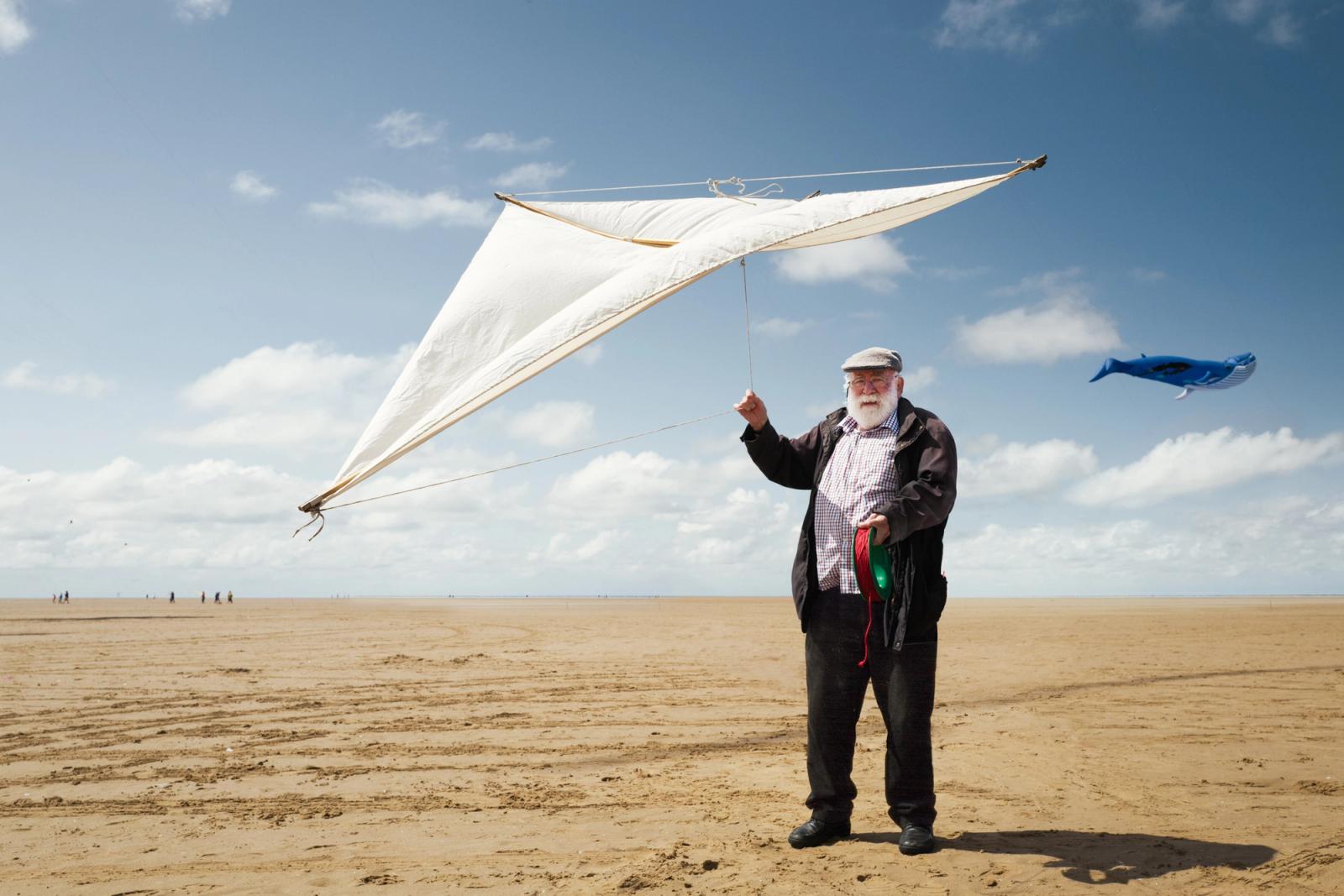 A man on a beach, flying a large white kite with a red and blue tail, in the background a blue kite is visible, flying high in the sky.