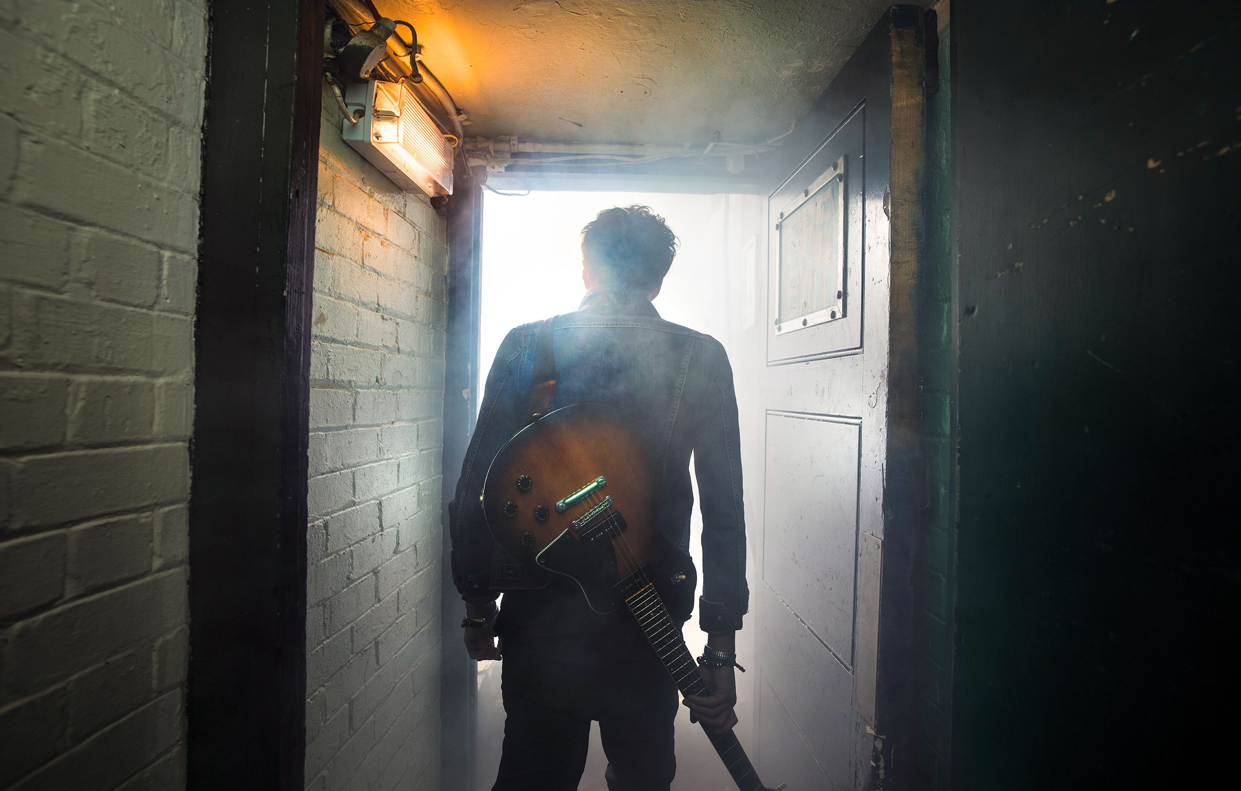 A man in a dark denim jacket and jeans, holding a guitar and standing in a dimly lit, industrial-style hallway with a door at the end, and a brick wall to the left.