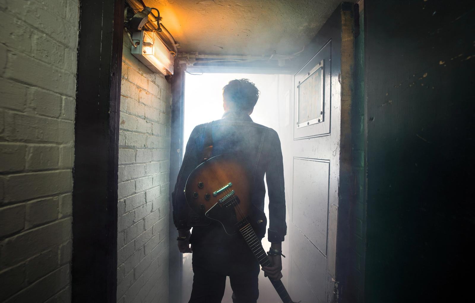 A man in a dark denim jacket and jeans, holding a guitar and standing in a dimly lit, industrial-style hallway with a door at the end, and a brick wall to the left.