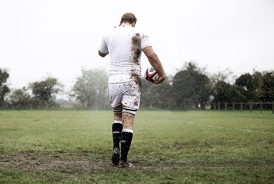 A man in a white rugby jersey and shorts, covered in mud, walking away from the camera, holding a rugby ball.