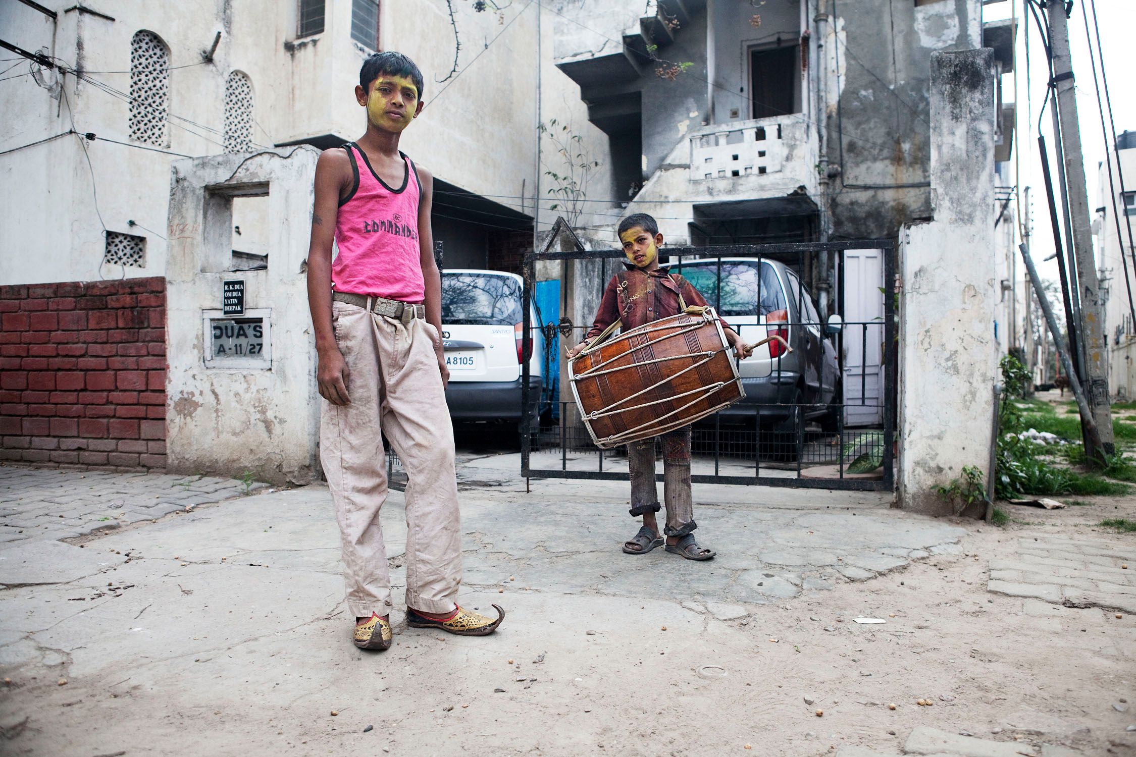 Two young boys with yellow face paint standing in front of a white building with a black gate and a white van behind them.