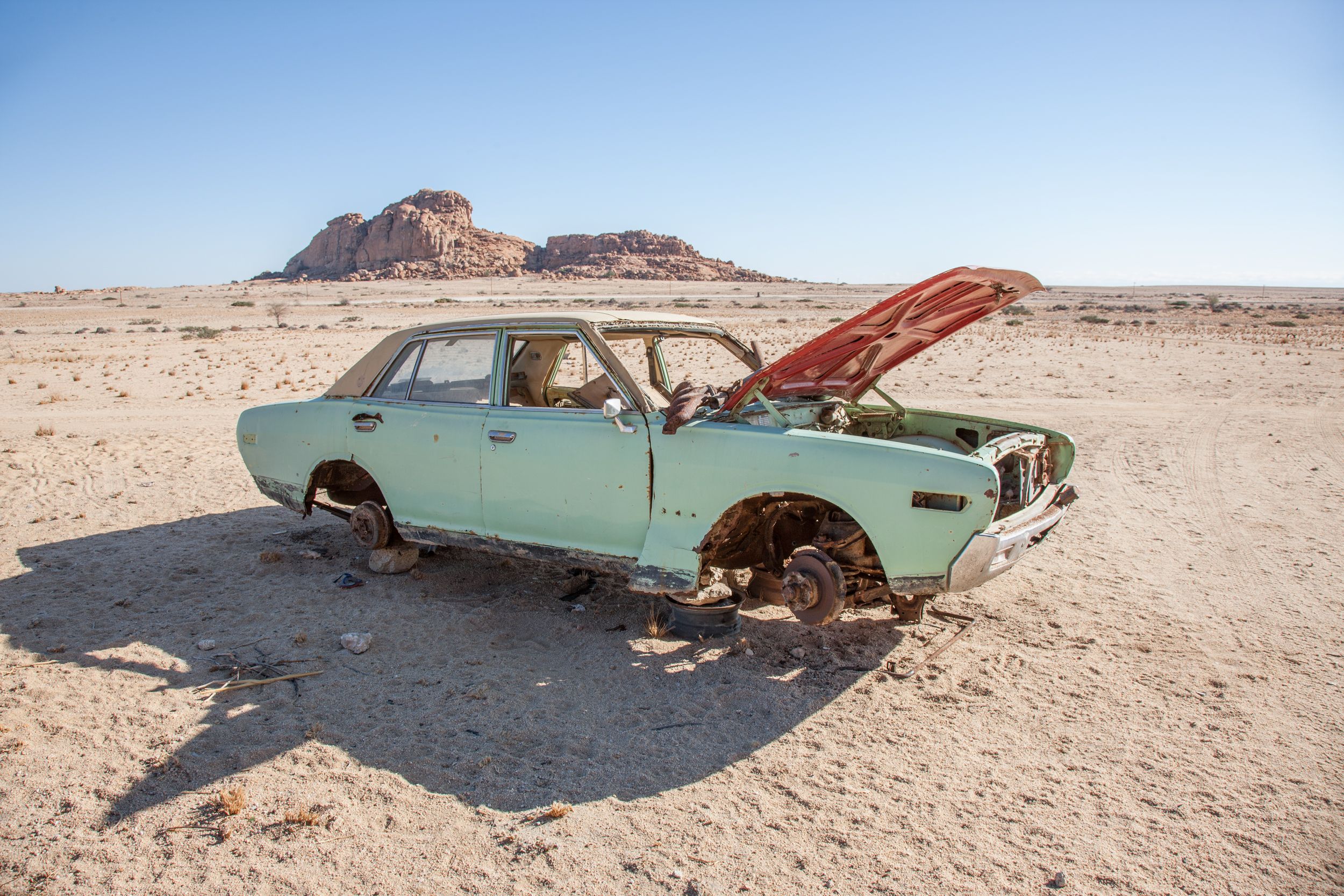 A car, likely a 1960s or 1970s model, is abandoned in the desert, with its engine and wheels removed, and its body is rusted and damaged, with a broken windshield and a flat tire.
