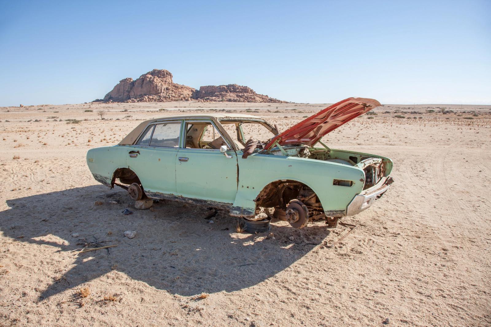 A car, likely a 1960s or 1970s model, is abandoned in the desert, with its engine and wheels removed, and its body is rusted and damaged, with a broken windshield and a flat tire.