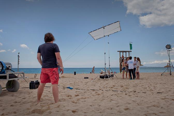 A man in a black t-shirt and red shorts standing on a beach, looking out at a camera crew and a large lighting rig.