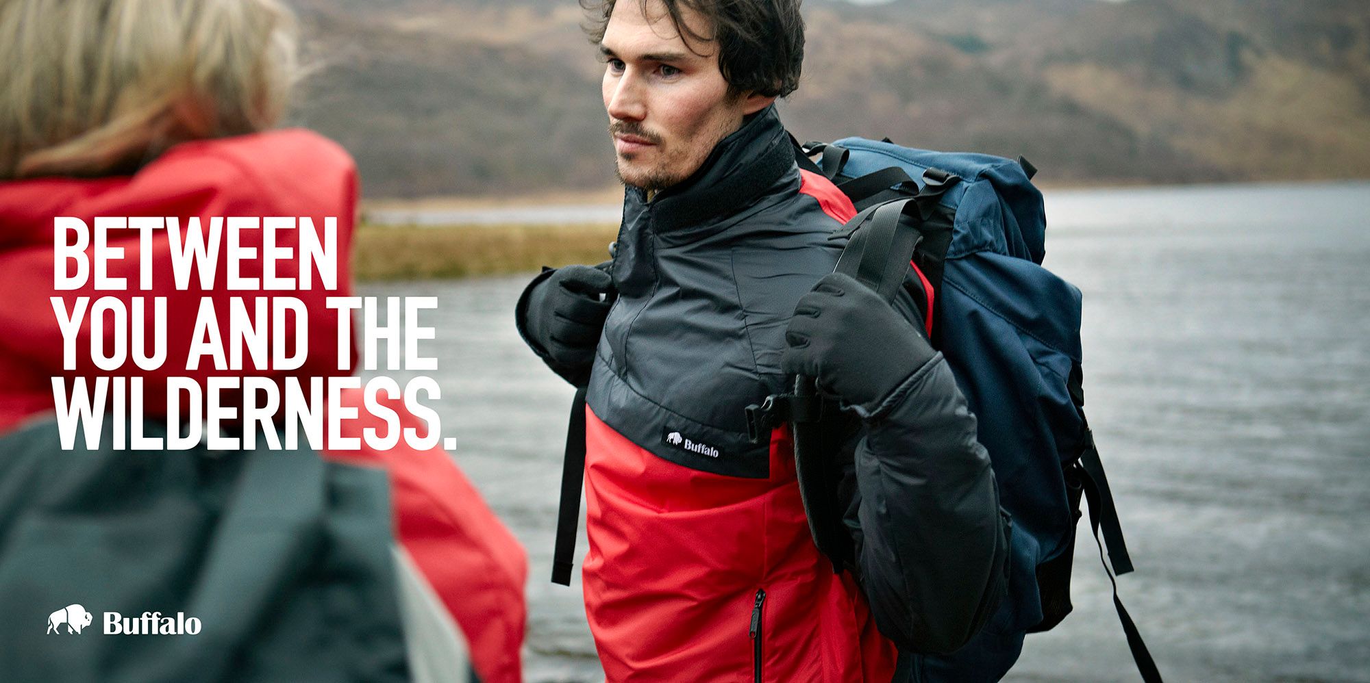 A man in a red and black jacket with a backpack on his back, standing in front of a body of water with a woman in the background.