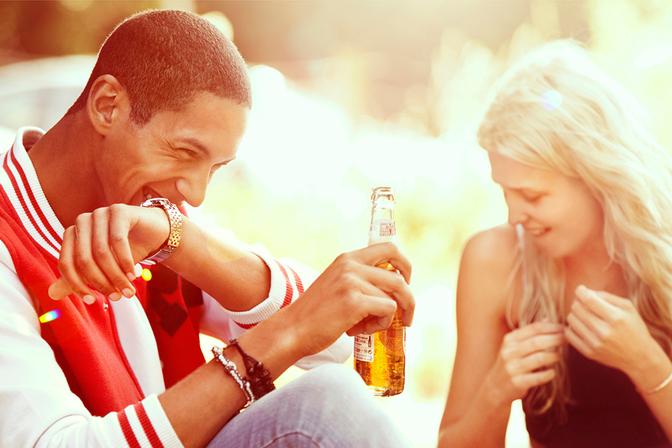A young woman with long blonde hair and a black tank top is laughing and holding a beer bottle with a man wearing a red and white striped rugby jersey and a gold watch.