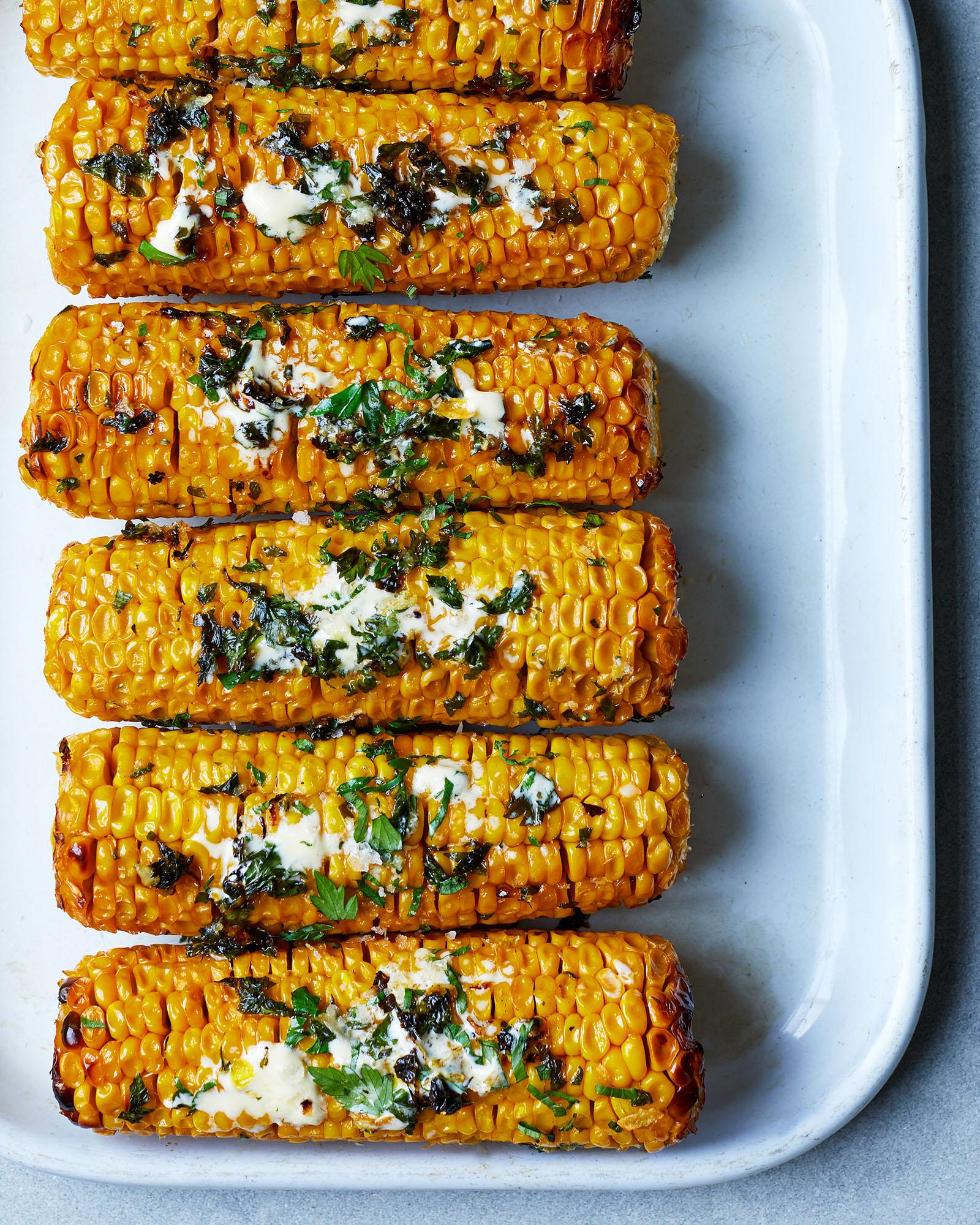 A close-up of five grilled corn on the cob, each topped with a pat of butter and a sprinkle of herbs.
