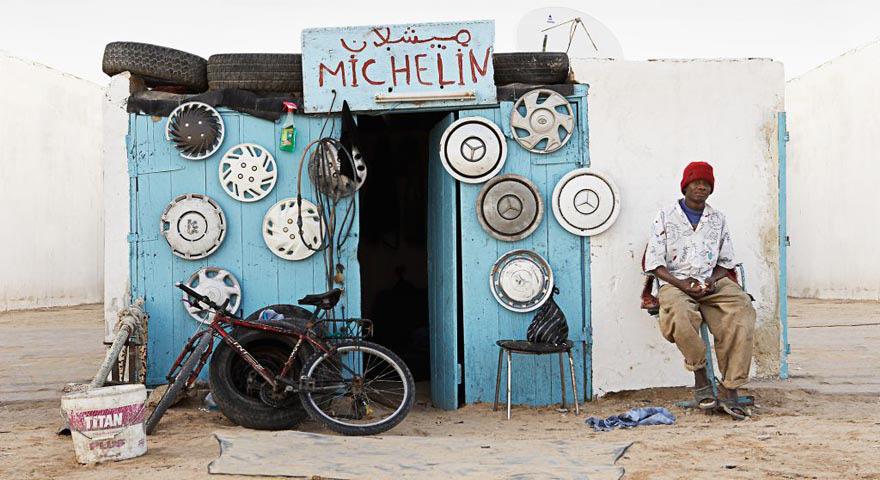A man sits in front of a blue and white building with an Arabic sign reading ‘Michel’, surrounded by a bicycle and other objects.