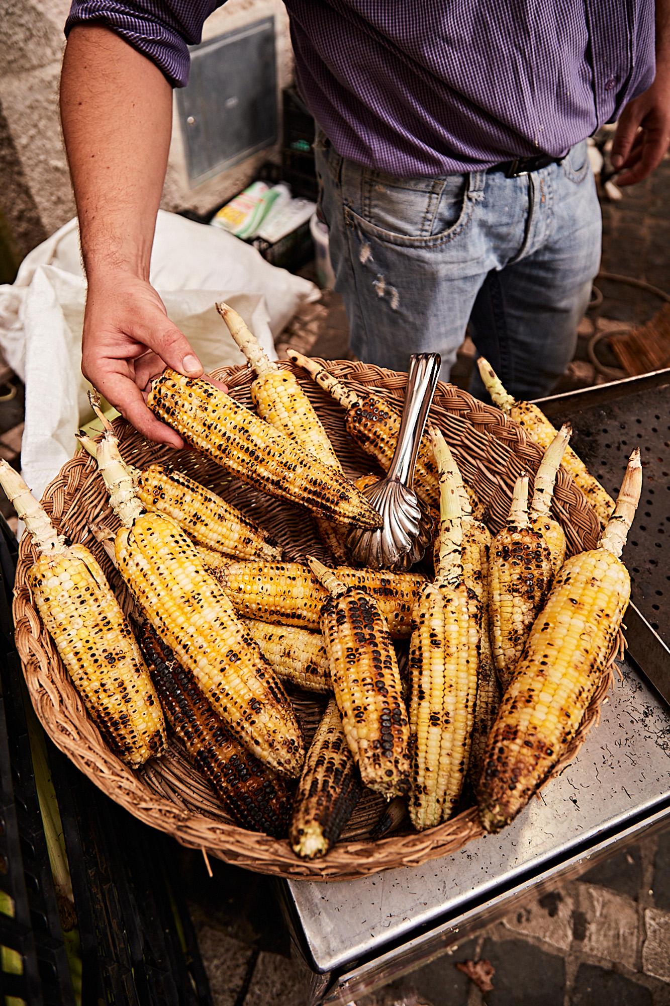 A person holding a basket of grilled corn on the cob, with a few pieces of corn on the cob in the basket and a few on the ground nearby.