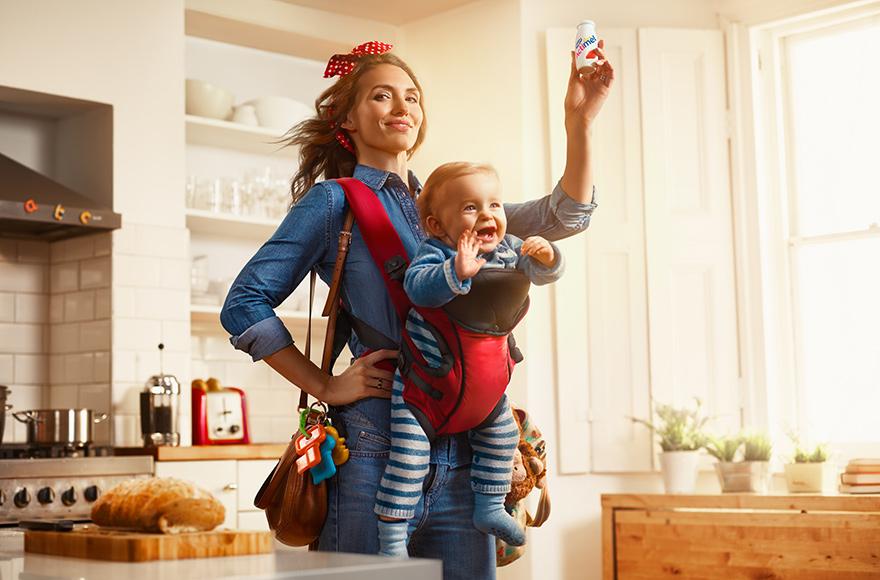 A woman wearing a denim shirt and jeans, holding a baby in a red baby carrier, in a modern kitchen.