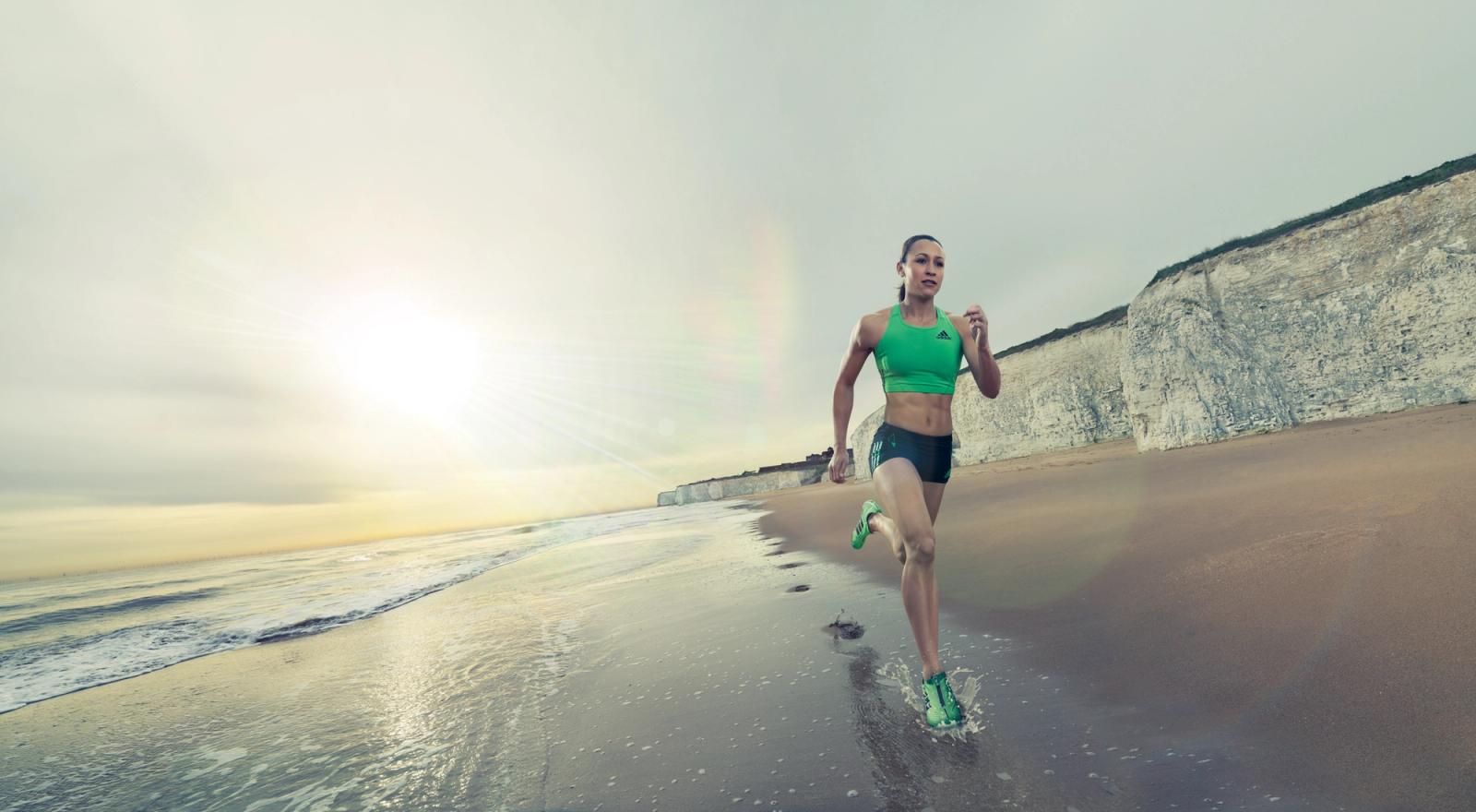 A woman in a green sports bra and black shorts running on a beach with a cliff in the background.