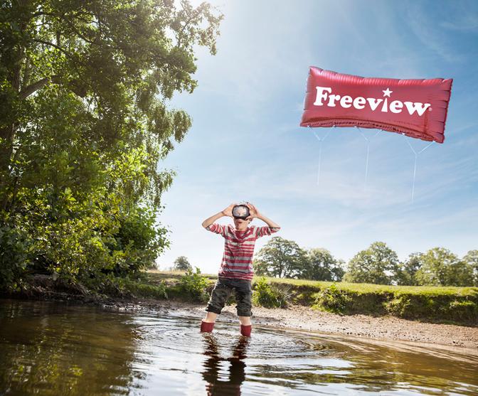 A young boy in a red and white striped shirt, holding a camera, standing in a shallow river, surrounded by trees and greenery.