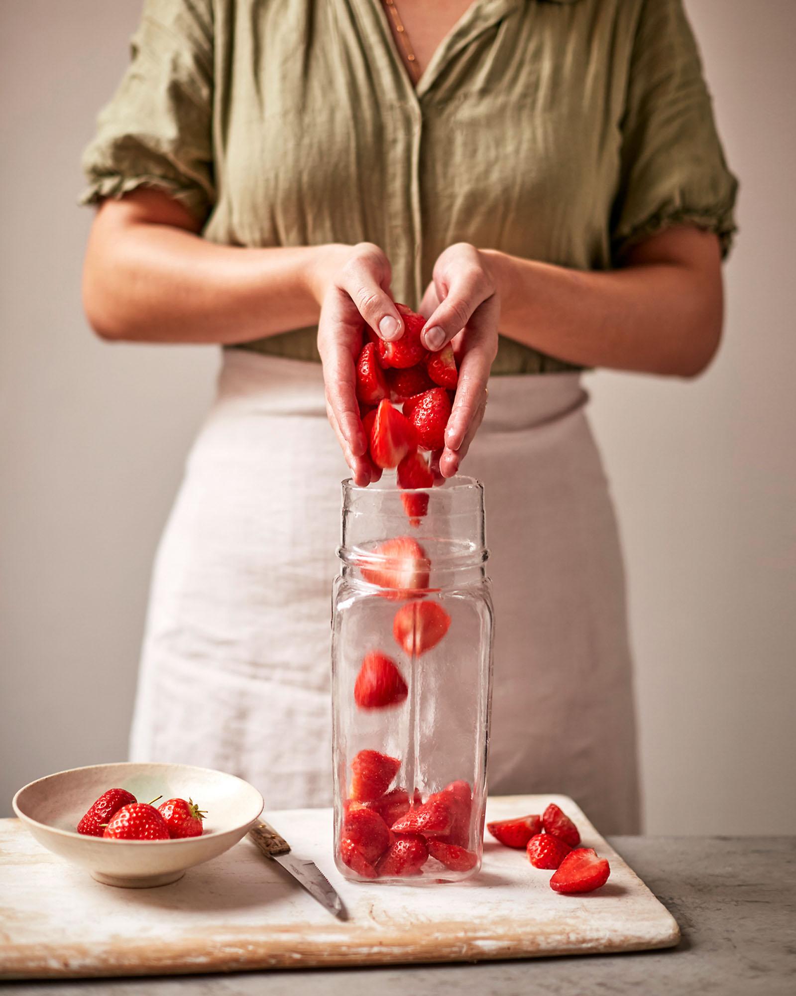 A woman in a green shirt and white apron is pouring strawberries from a glass jar into a bowl.
