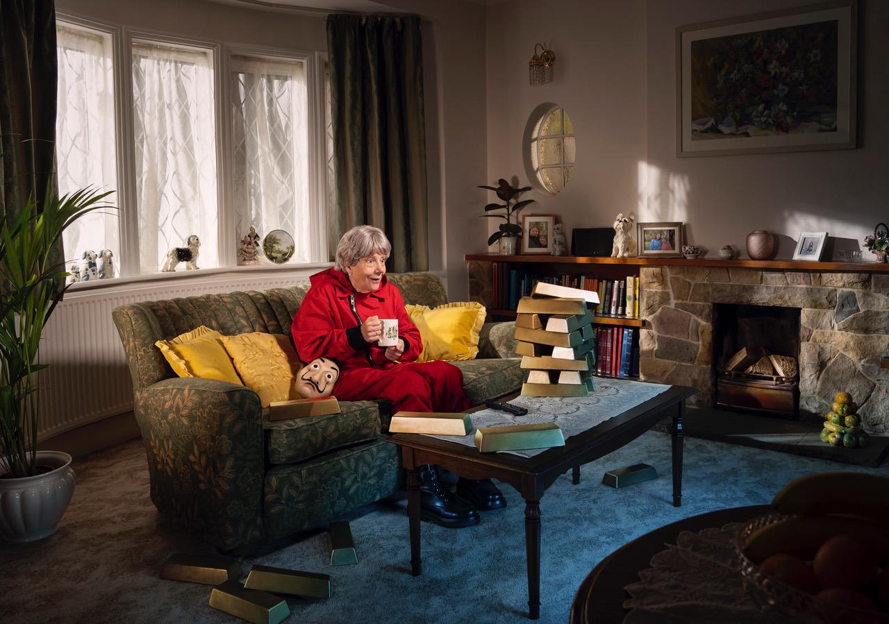 An older woman sits on a couch in a living room, surrounded by gold bars and a Jenga game.