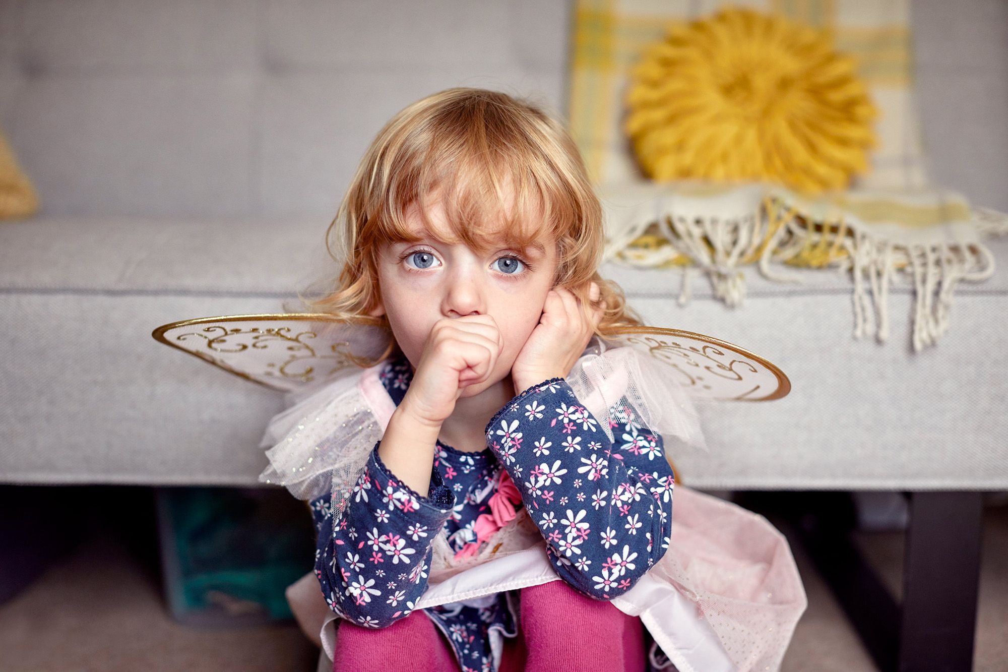 A young girl with blonde hair and blue eyes, wearing a fairy costume with wings and a tutu, sitting on a couch with a concerned expression.