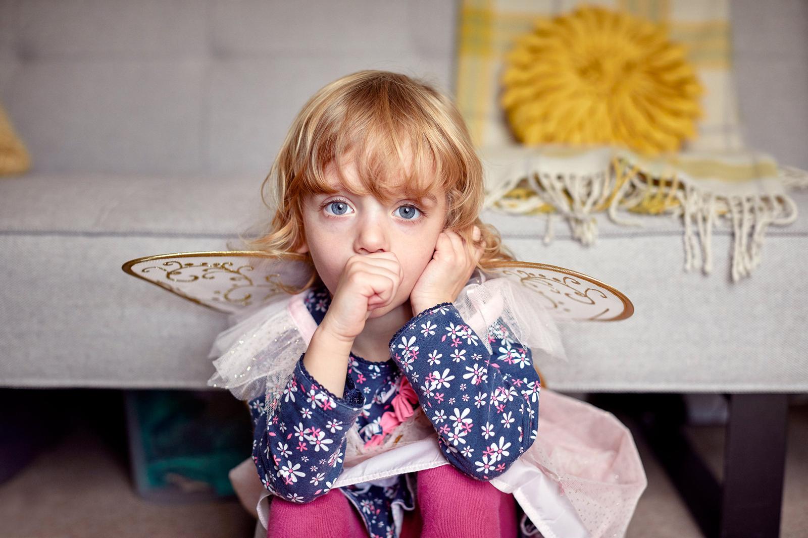 A young girl with blonde hair and blue eyes, wearing a fairy costume with wings and a tutu, sitting on a couch with a concerned expression.
