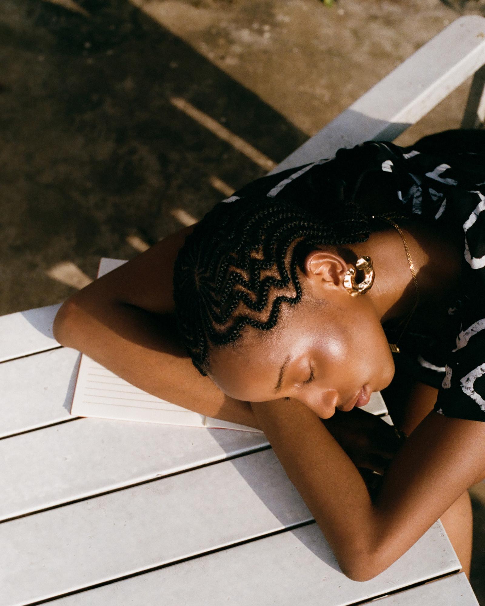 A young woman with braided hair and a black and white patterned shirt is sleeping on a white table, surrounded by books and papers.