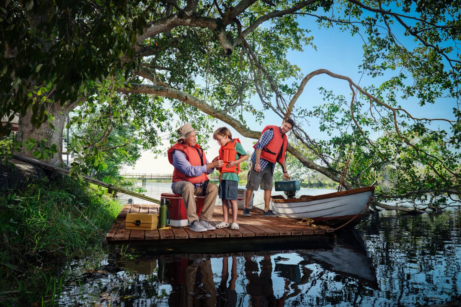 A man, a boy, and a man on a small wooden boat on a lake, all wearing life jackets.