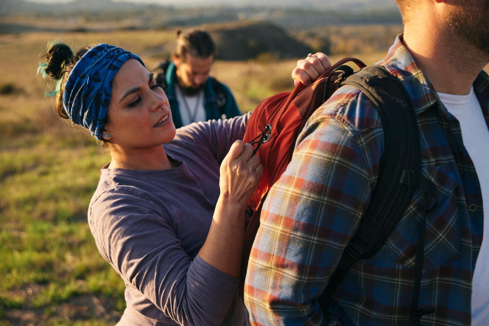 A woman with a blue bandana on her head is helping a man with a red backpack on his shoulders, both in a field. The woman is adjusting the straps of the backpack.