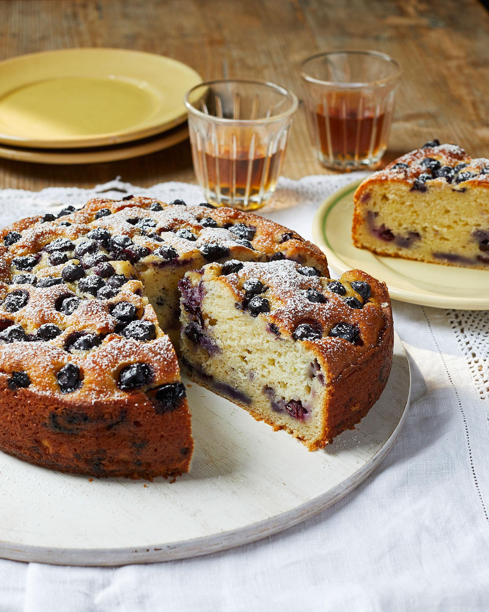 A golden-brown cake with blueberries and powdered sugar, displayed on a white plate with a slice missing.