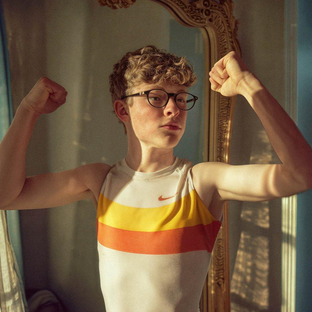 A young man flexes his arms in front of a large gold-framed mirror.