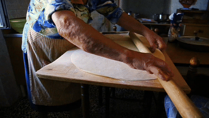 This image shows an older woman in an apron, rolling out dough on a wooden board with a rolling pin. She is wearing a blue and white patterned top and a brown and white checked.
